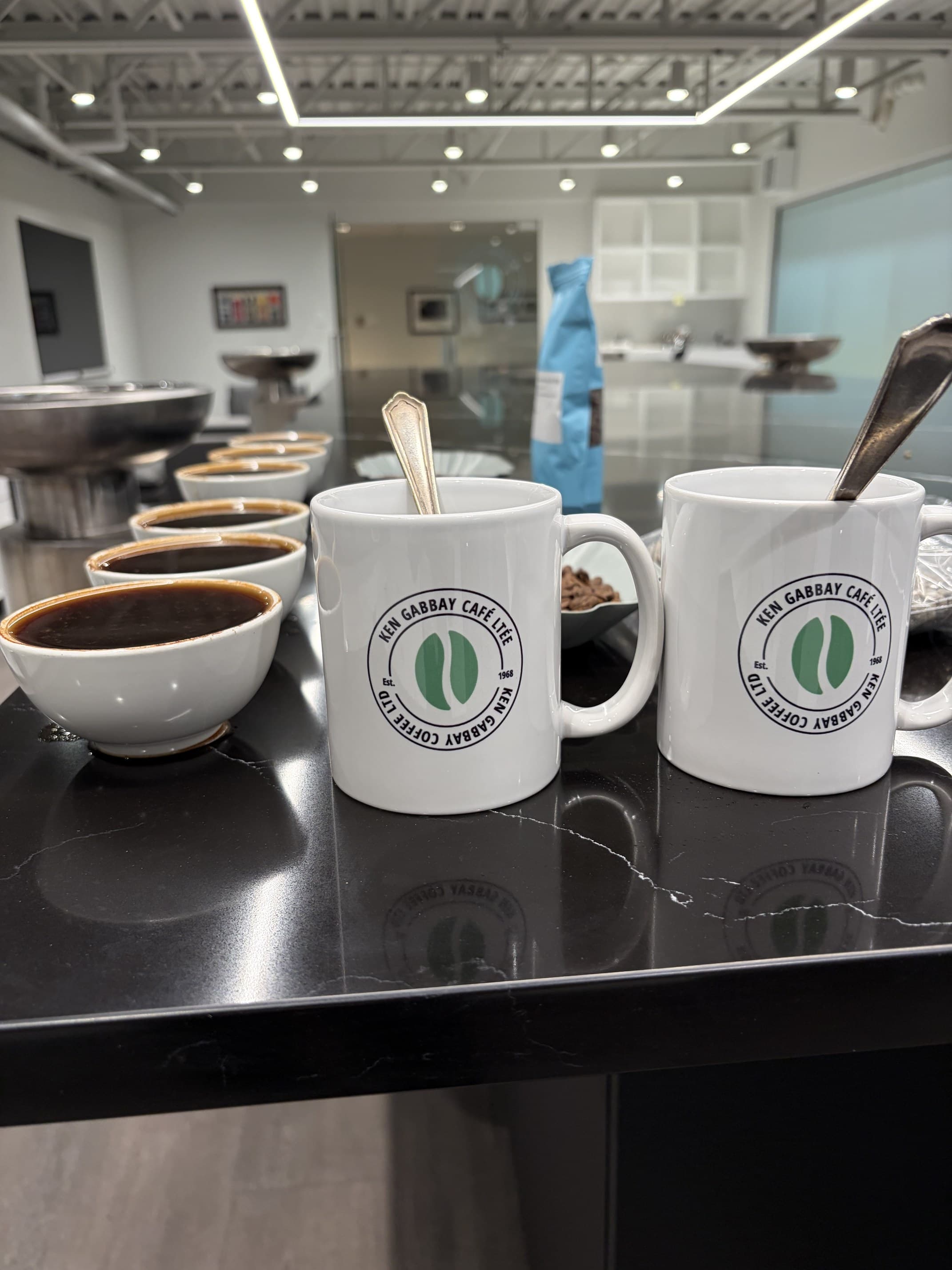 A row of white cupping bowls filled with brewed coffee along a black stone table, with two mugs printed with the Ken Gabbay Coffee seal in the foreground and spoons resting in them.