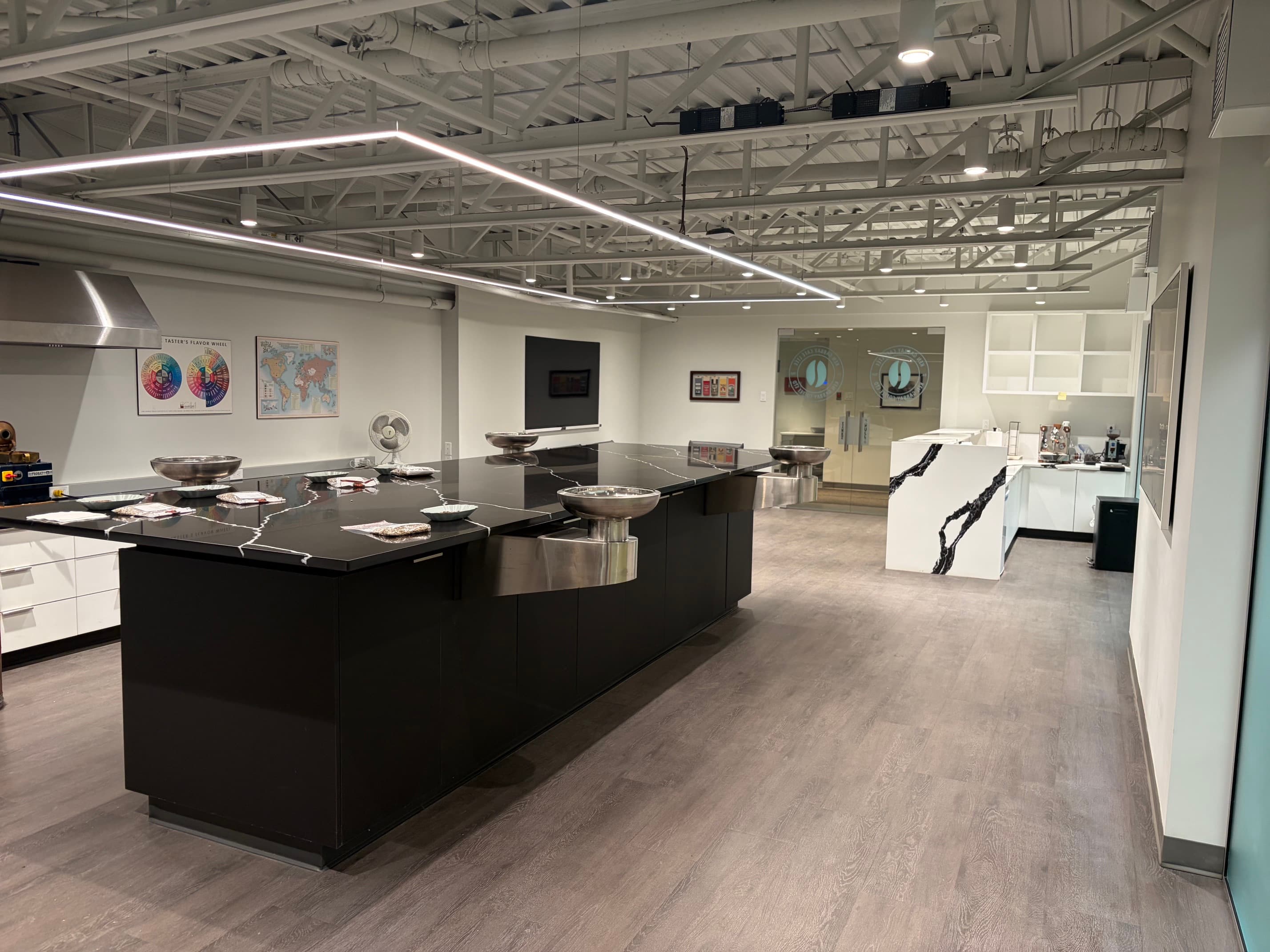 The long black cupping table with steel bowls and sample trays set along it, a taster's flavor wheel and a world coffee map on the wall behind.
