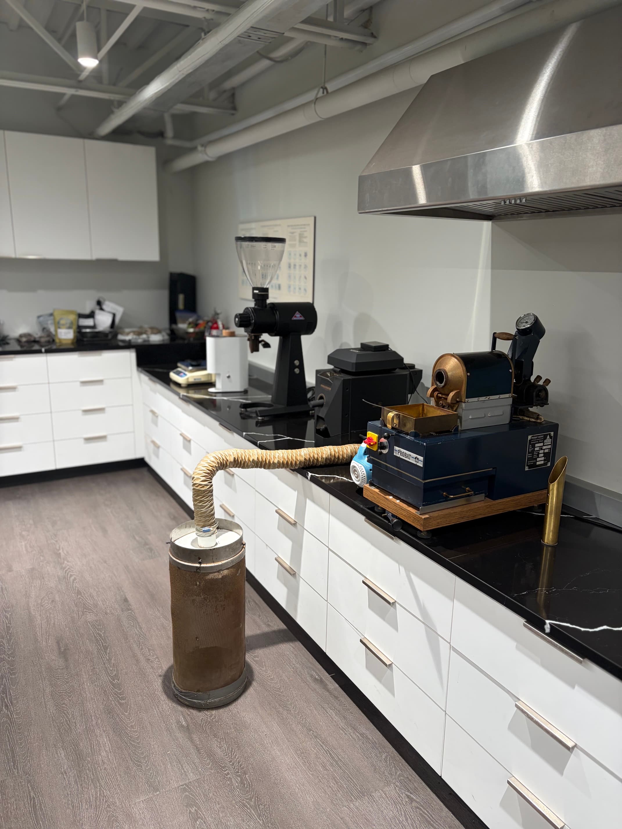 A blue Probat sample roaster on the bench, a duct running from it down into a metal bin on the floor, a grinder alongside.