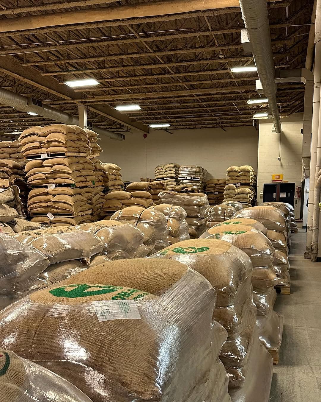 An aisle of the Montreal warehouse, wrapped pallets of green coffee in the foreground and jute bags stacked high behind them.
