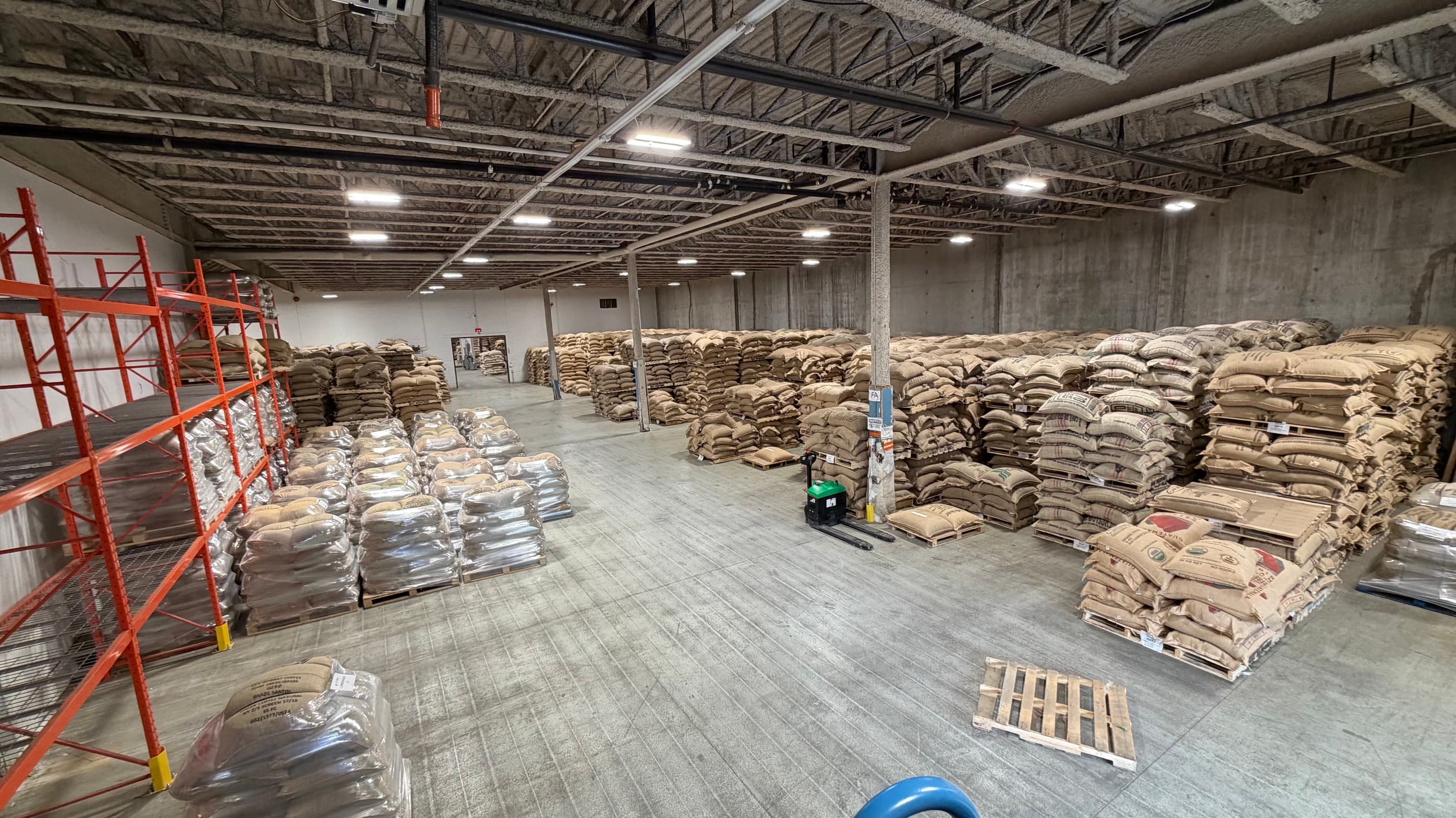 A wide view across the warehouse floor, wrapped pallets and stacked jute bags on both sides and an electric pallet jack parked in the middle.
